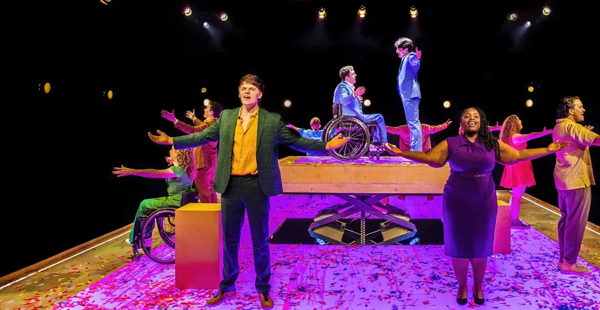 Production photo. Cast stood with open arms in colourful lighting. 