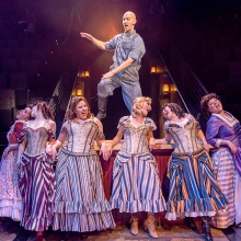 Production photo from The Unsinkable Molly Brown. A man dances on a table with a group of women singing in front of him
